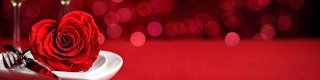 Et hjerteformet rødt rose ligger på en hvid tallerken med bestik ved siden af. I baggrunden ses to glassene fyldt med gylden drink, alt på en rød baggrund. Billedet giver en romantisk stemning.